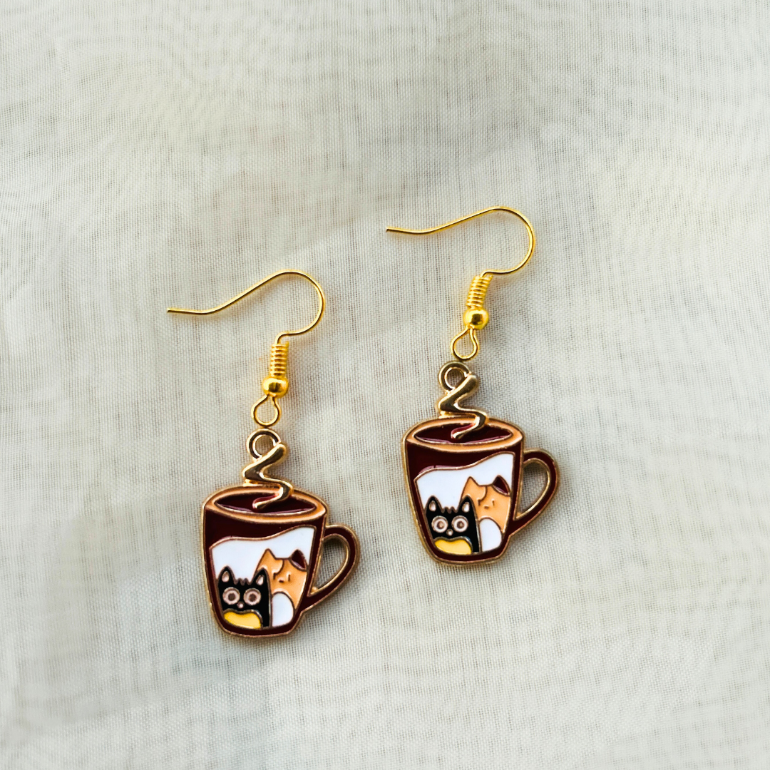 A pair of earrings featuring metal enamel charms with cat illustrations inside cups, attached to gold-colored earring hangings.