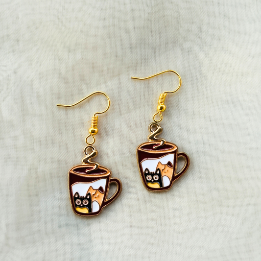 A pair of earrings featuring metal enamel charms with cat illustrations inside cups, attached to gold-colored earring hangings.